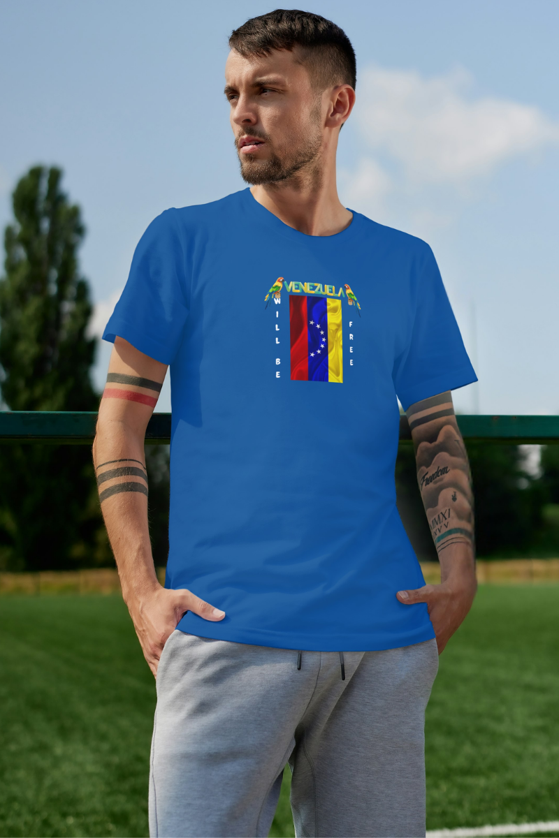 A young man is wearing a blue T-shirt featuring the 7-star Venezuelan flag, the name VENEZUELA at the top, and one macaw on each side. The words WILL BE and FREE are all placed vertically next to the flag. 
He’s standing, looking to his right.