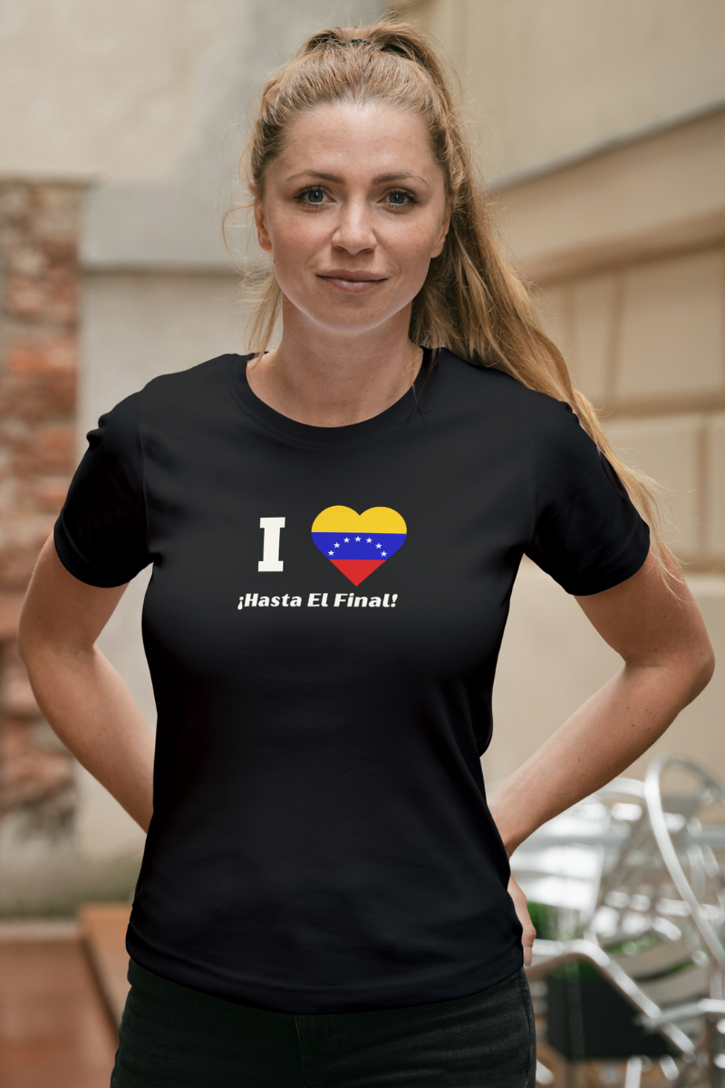 A blonde woman wears a black T-shirt featuring the 7-star Venezuelan flag in the shape of a heart, the word "I" on the left, and the text "¡HASTA EL FINAL!" below.
She’s standing, facing forward.