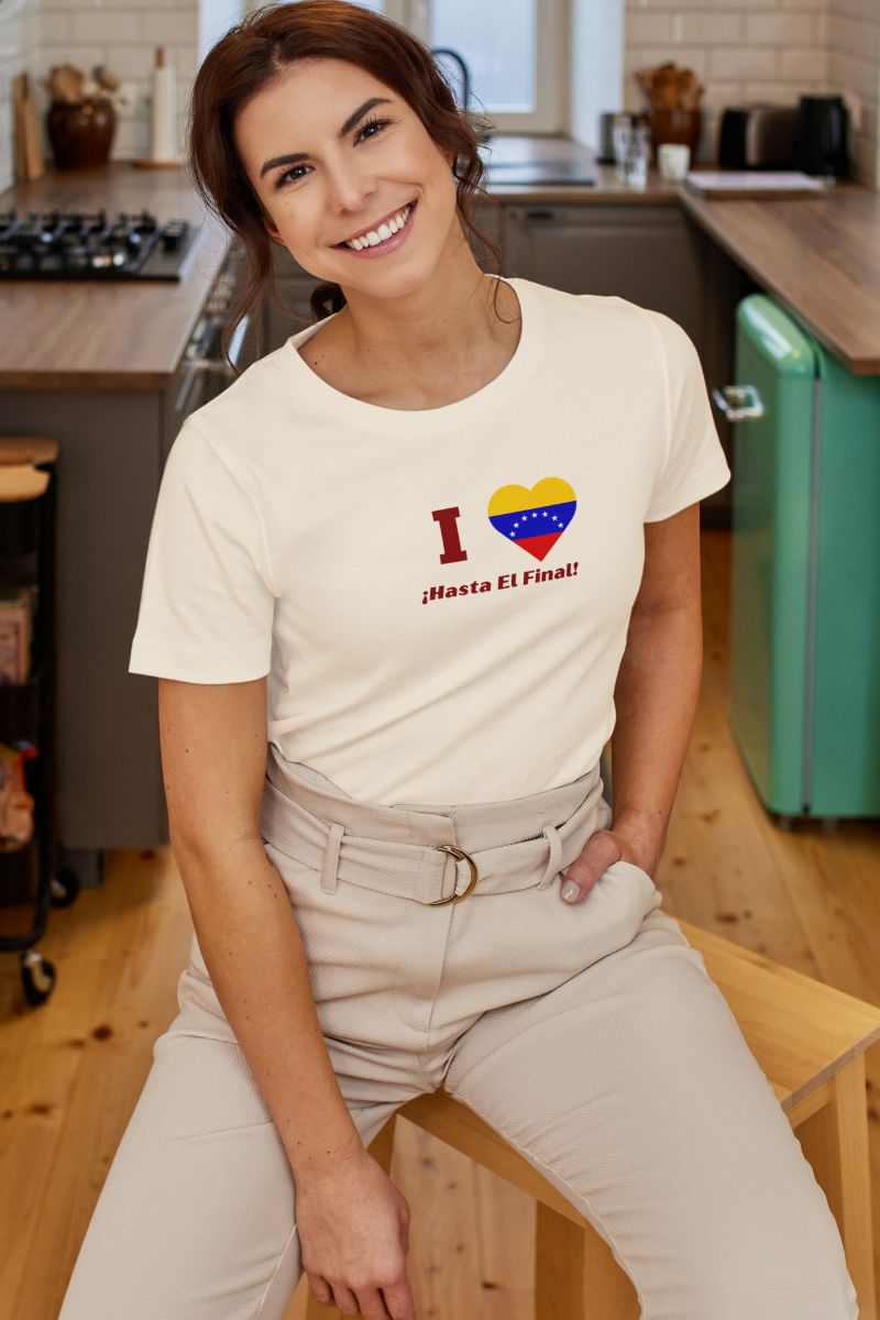 A smiling woman wears a pale orange T-shirt featuring the 7-star Venezuelan flag in the shape of a heart, the word "I" on the left, and the text "¡HASTA EL FINAL!" below.
She’s in a kitchen, sitting on a small wooden bench.