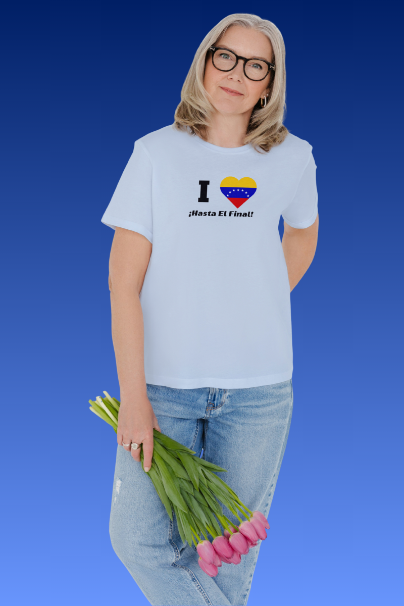 A blonde woman wears a light blue T-shirt featuring the 7-star Venezuelan flag in the shape of a heart, the word "I" on the left, and the text "¡HASTA EL FINAL!" below.
She’s facing forward, holding some flowers with her right hand.