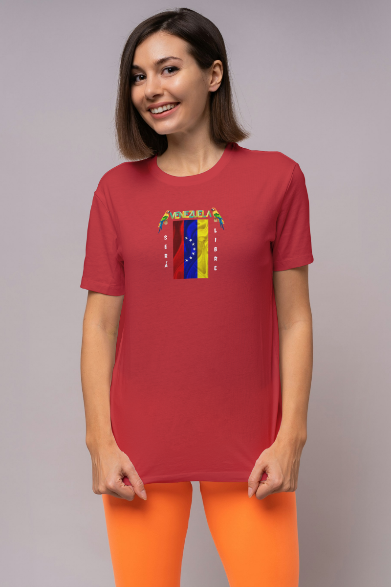 Una mujer joven y sonriente viste una franela roja con la bandera venezolana de 7 estrellas, el nombre VENEZUELA arriba y una guacamaya a cada lado. Las palabras SERÁ y LIBRE se ubican verticalmente junto a la bandera.