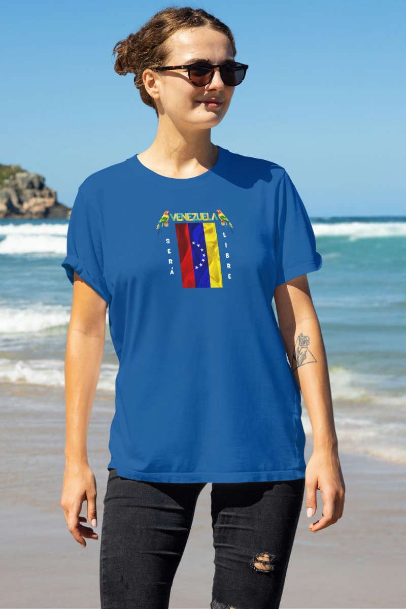 Una mujer sonriente, en una playa, viste una franela azul con la bandera venezolana de 7 estrellas, el nombre VENEZUELA arriba y una guacamaya a cada lado. Las palabras SERÁ y LIBRE se ubican verticalmente junto a la bandera. 