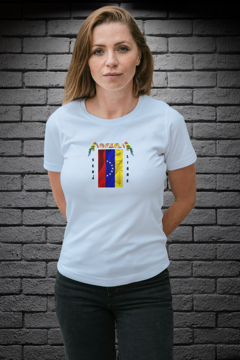 Una mujer joven, de pie y con ambas manos detrás de su espalda, viste una franela azul claro con la bandera venezolana de 7 estrellas, el nombre VENEZUELA arriba y una guacamaya a cada lado. Las palabras SERÁ y LIBRE se ubican verticalmente junto a la bandera.
