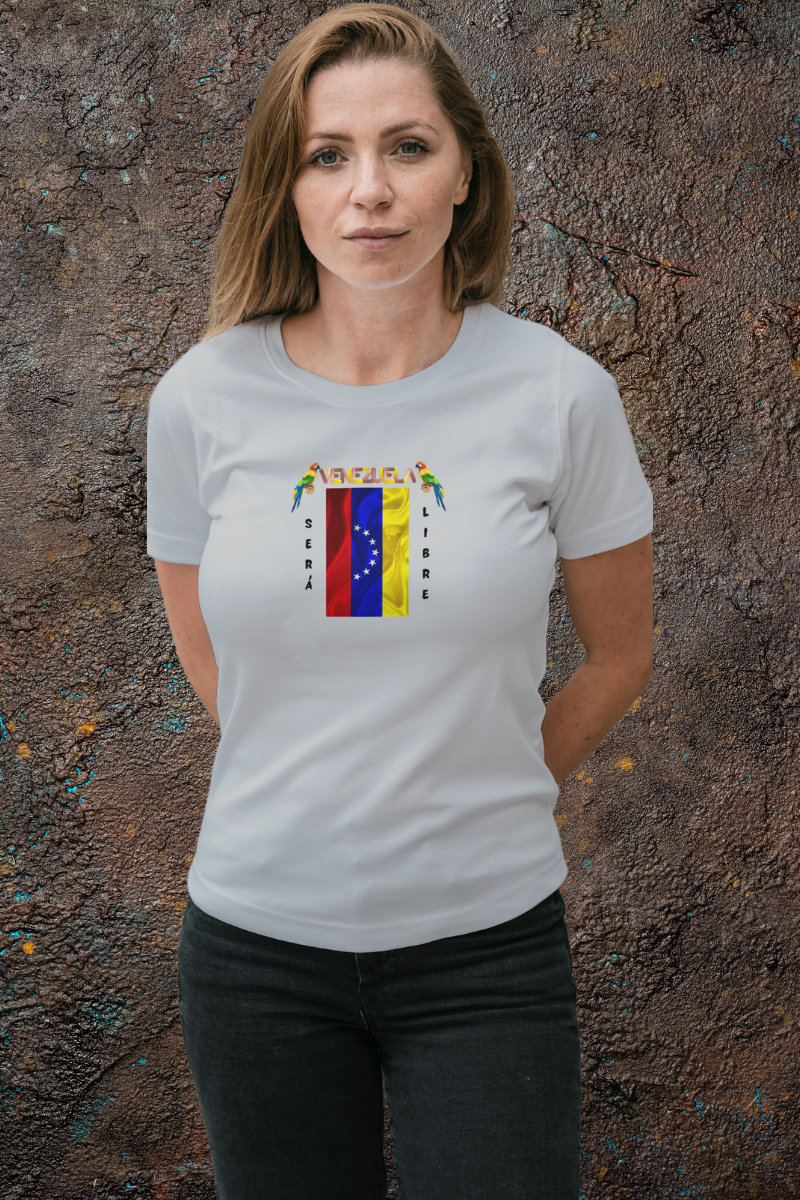 Una mujer joven, de pie y con ambas manos detrás de su espalda, viste una franela gris claro con la bandera venezolana de 7 estrellas, el nombre VENEZUELA arriba y una guacamaya a cada lado. Las palabras SERÁ y LIBRE se ubican verticalmente junto a la bandera.