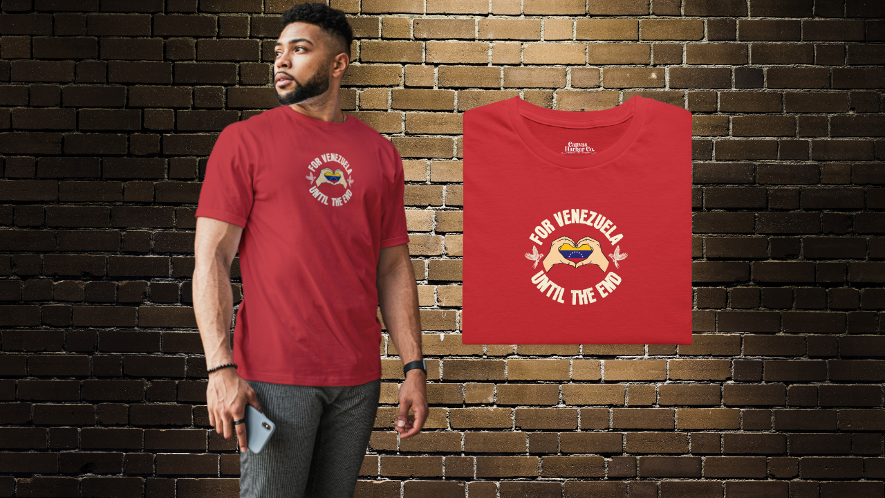 Red folded T-shirt featuring the Venezuelan flag inside heart-shaped hands, the text “FOR VENEZUELA UNTIL THE END” around it, and one macaw on each side.
On the left, an Afro-American man wears the T-shirt. He’s standing, looking to his right.