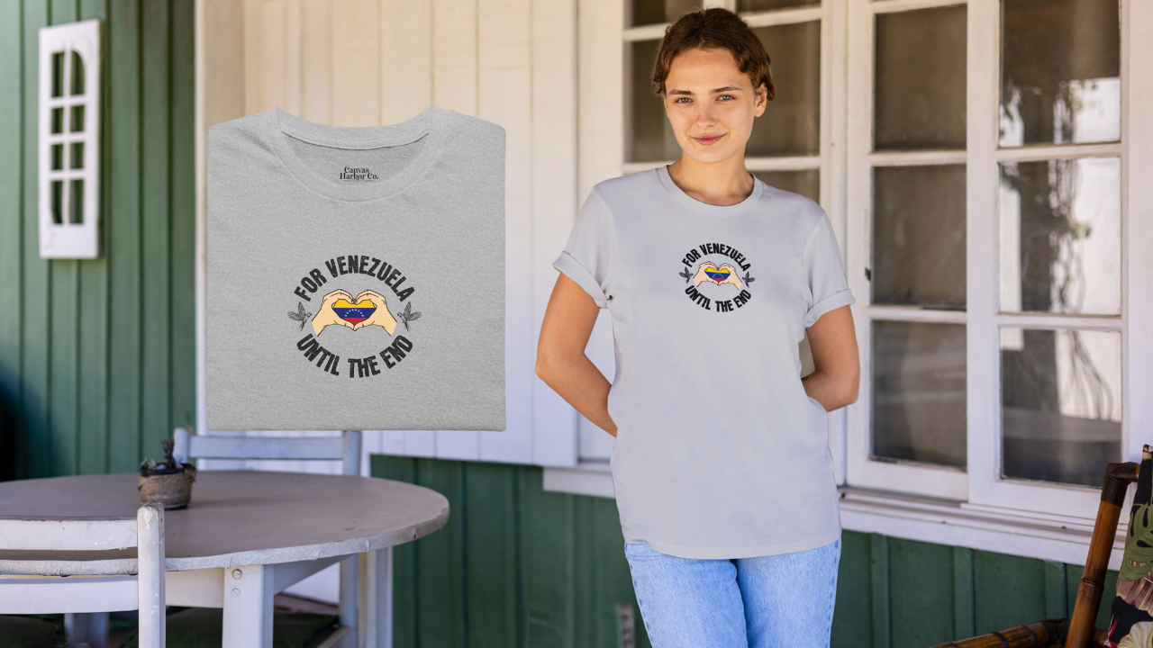 Light gray folded T-shirt featuring the Venezuelan flag inside heart-shaped hands, the text “FOR VENEZUELA UNTIL THE END” around it, and one macaw on each side.
On the right, a young woman wears the T-shirt. She’s smiling, facing forward, holding both hands behind her back.