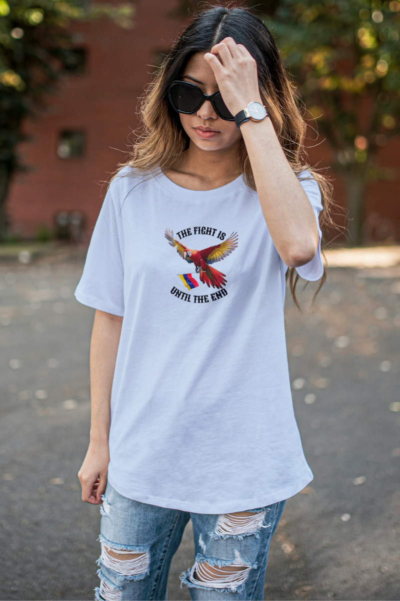 A young woman wears a light blue T-shirt featuring an image of a macaw and the Venezuelan flag. The text "THE FIGHT IS UNTIL THE END" surrounds the bird.
She’s standing, wearing sunglasses.