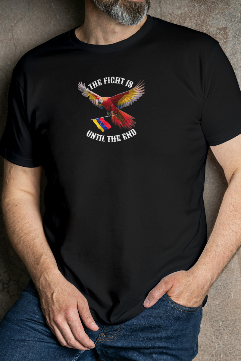 A man wears a black T-shirt featuring an image of a macaw and the Venezuelan flag. The text "THE FIGHT IS UNTIL THE END" surrounds the bird.
He’s standing, with his left hand in the pocket of his pants.