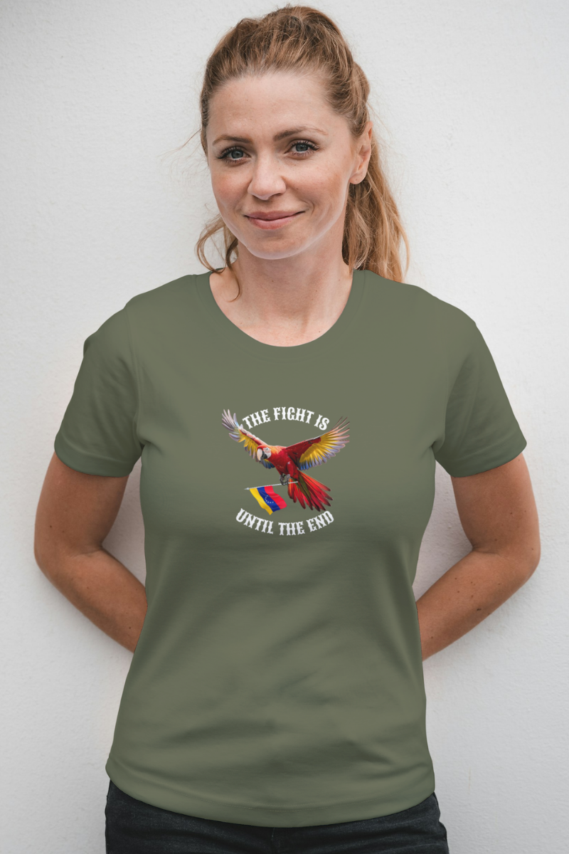A blonde smiling woman wears an olive-green T-shirt featuring an image of a macaw and the Venezuelan flag. The text "THE FIGHT IS UNTIL THE END" surrounds the bird.
She’s standing, holding her hands behind her back.