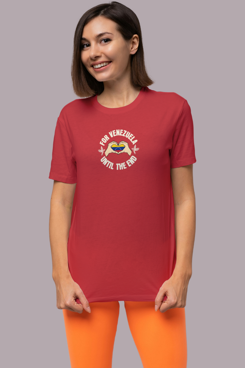 A young woman wears a red T-shirt featuring the Venezuelan flag inside heart-shaped hands, the text “FOR VENEZUELA UNTIL THE END” around it, and one macaw on each side.
She’s standing, smiling, facing forward.