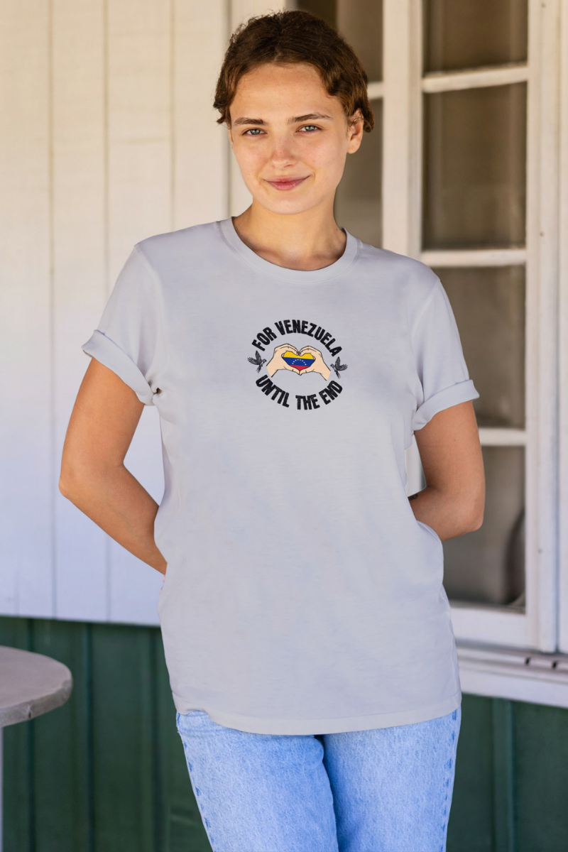 A young woman wears a light gray T-shirt featuring the Venezuelan flag inside heart-shaped hands, the text “FOR VENEZUELA UNTIL THE END” around it, and one macaw on each side.
She’s smiling, facing forward, holding both hands behind her back.