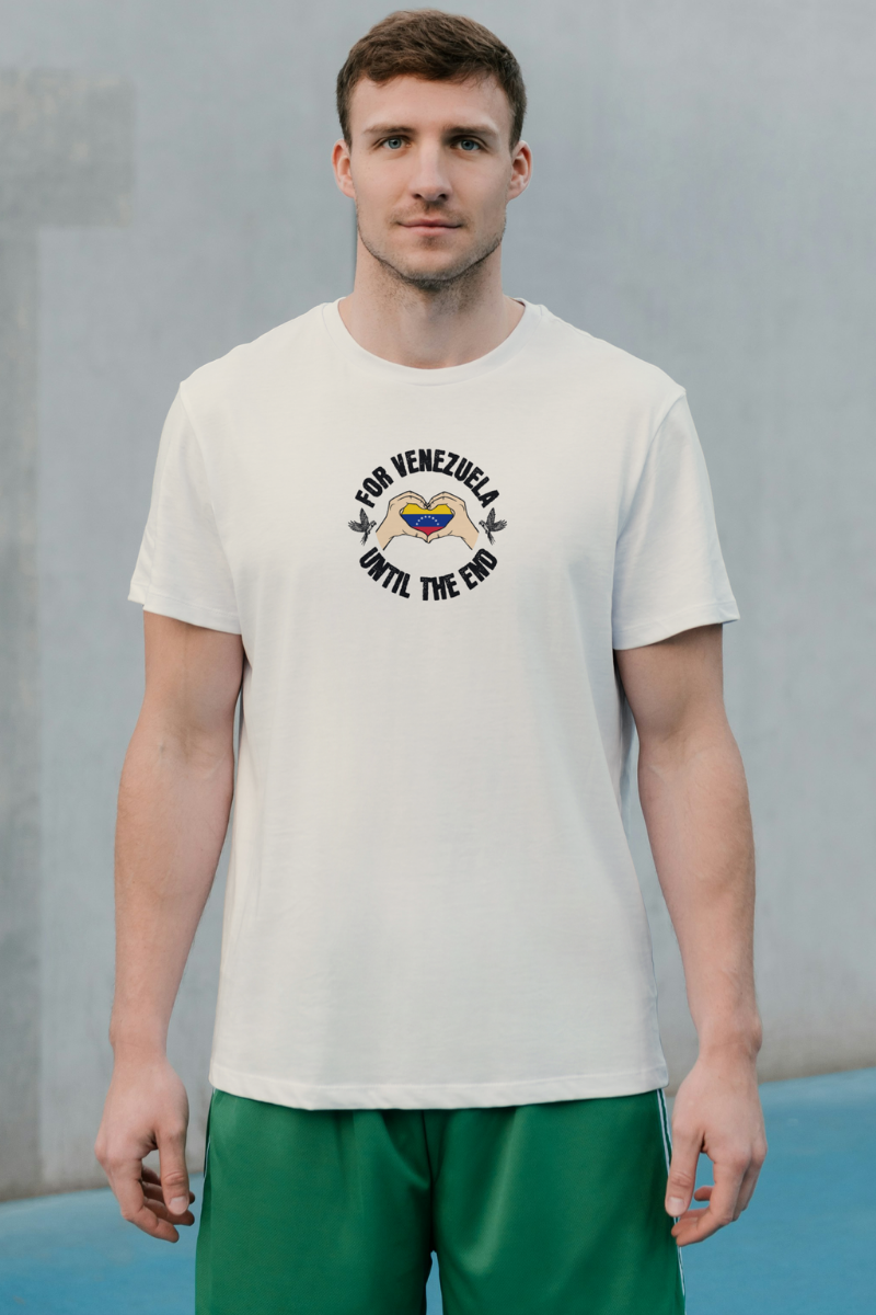 A young man wears a pale orange T-shirt featuring the Venezuelan flag inside heart-shaped hands, the text “FOR VENEZUELA UNTIL THE END” around it, and one macaw on each side.
He’s standing, facing forward.