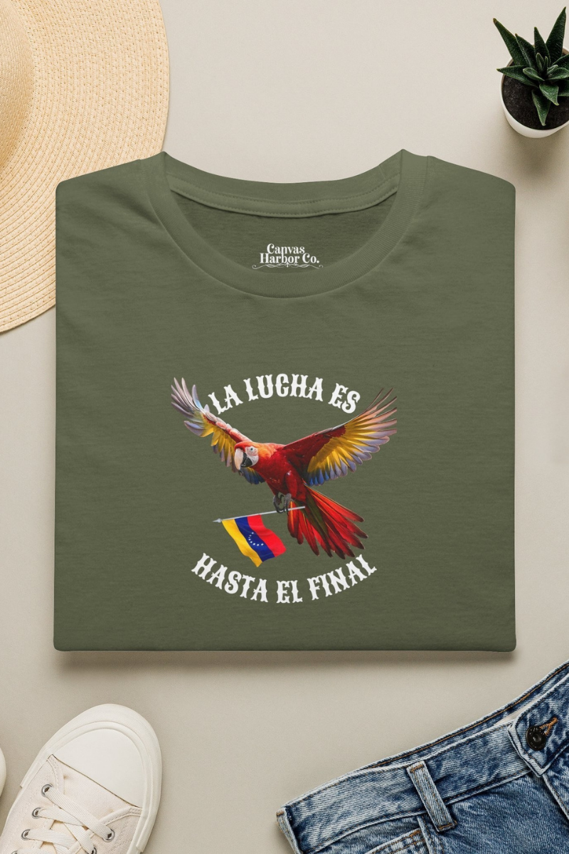 Franela verde oliva doblada sobre una mesa, con la imagen de un guacamayo y la bandera venezolana. La frase "LA LUCHA ES HASTA EL FINAL" rodea al ave.