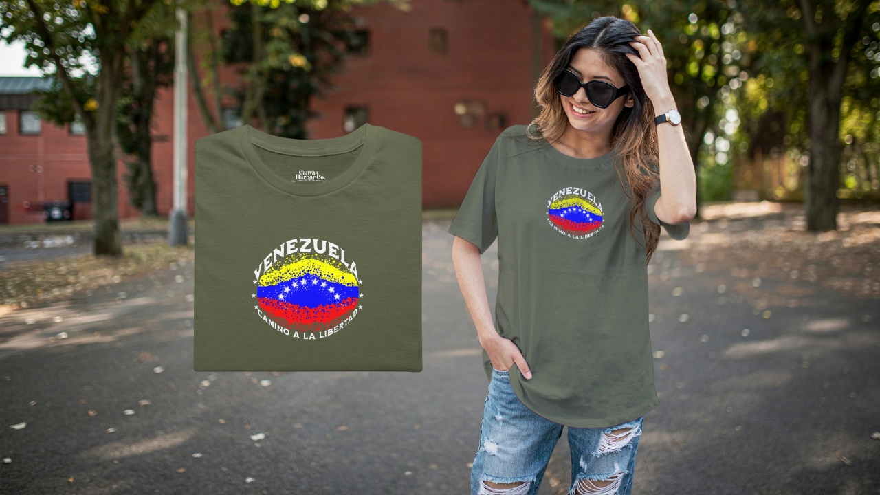 Franela color verde oliva, doblada, con una bandera venezolana de forma elíptica salpicada de puntos (como una explosión), con el nombre "VENEZUELA” arriba, el texto “CAMINO A LA LIBERTAD" abajo y 3 estrellas en cada lado.
A la derecha, una mujer joven y sonriente viste la franela. Ella está de pie y usa lentes de sol.