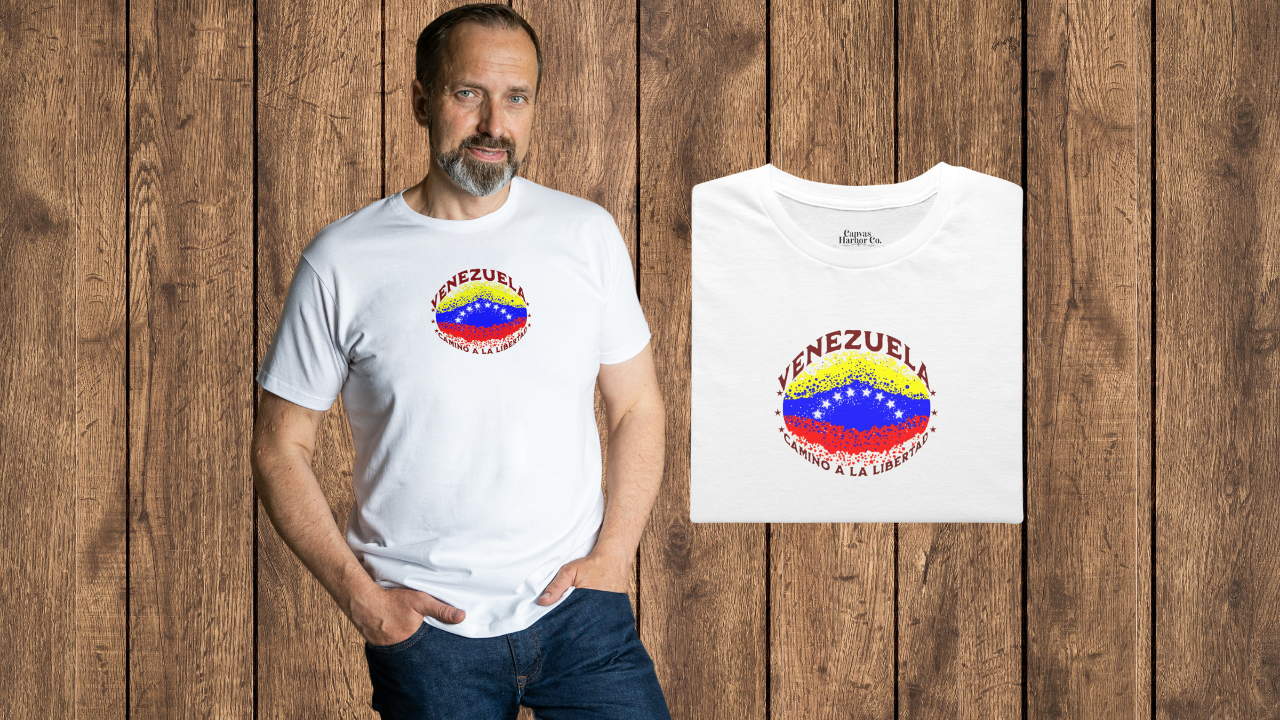 Franela blanca, doblada, con una bandera venezolana de forma elíptica salpicada de puntos (como una explosión), con el nombre "VENEZUELA” arriba, el texto “CAMINO A LA LIBERTAD" abajo y 3 estrellas en cada lado.
A la izquierda, un hombre viste la franela. Él está de pie, mirando al frente, con ambas manos en los bolsillos de su pantalón.