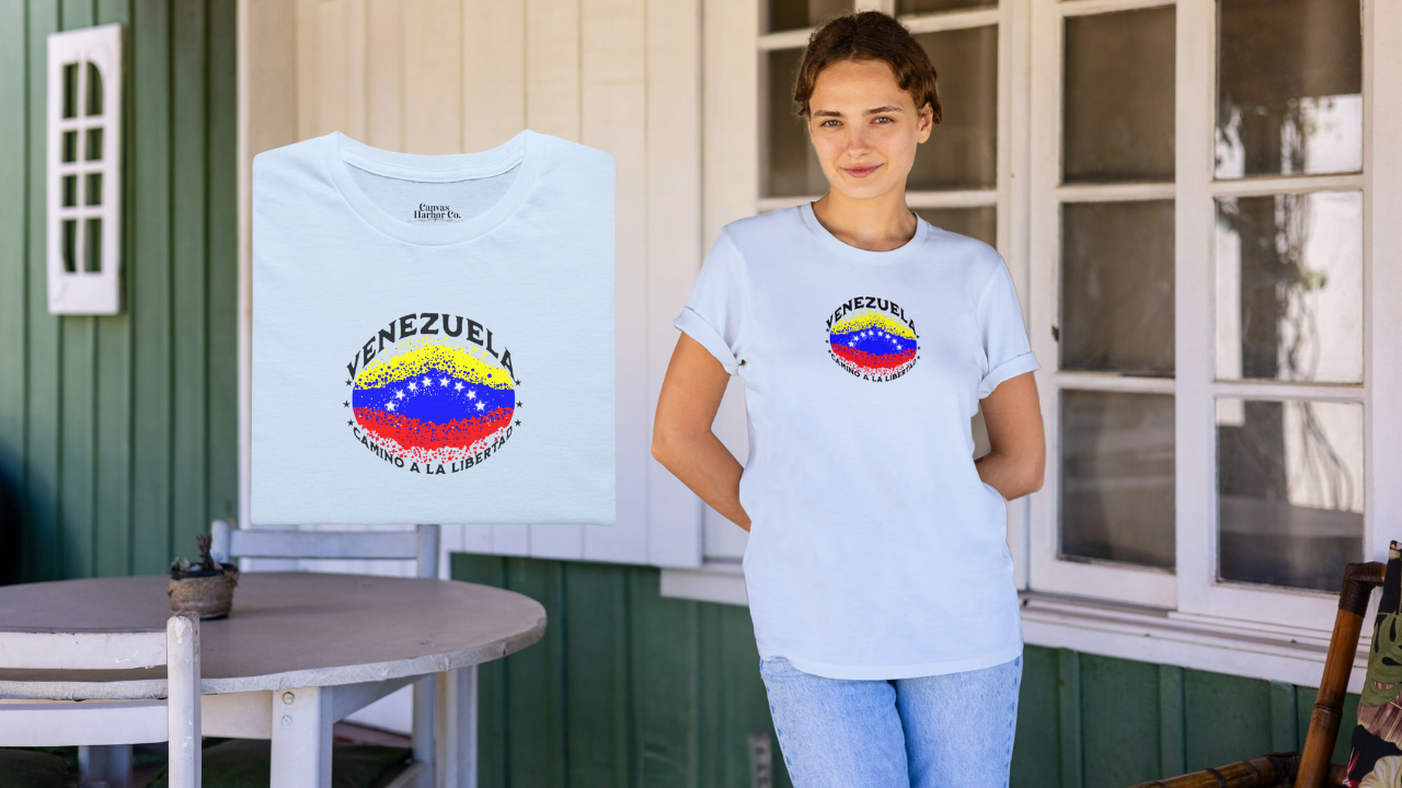 Franela color azul claro, doblada, con una bandera venezolana de forma elíptica salpicada de puntos (como una explosión), con el nombre "VENEZUELA” arriba, el texto “CAMINO A LA LIBERTAD" abajo y 3 estrellas en cada lado.
A la derecha, una mujer joven viste la franela. Ella sonríe, mirando al frente, con ambas manos detrás de su espalda.