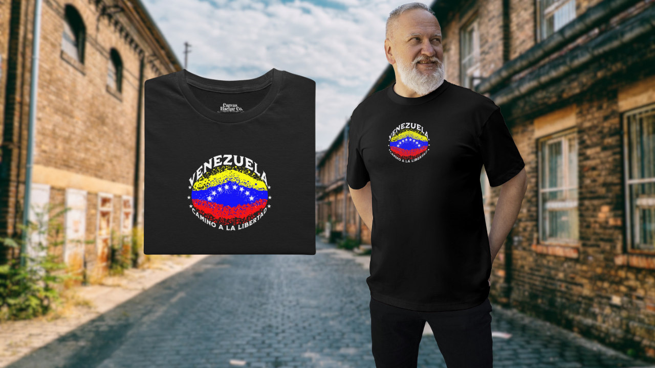 Franela negra, doblada, con una bandera venezolana de forma elíptica salpicada de puntos (como una explosión), con el nombre "VENEZUELA” arriba, el texto “CAMINO A LA LIBERTAD" abajo y 3 estrellas en cada lado.
A la derecha, un hombre con barba viste la franela. Él está de pie, mirando a su izquierda, con ambas manos detrás de su espalda.