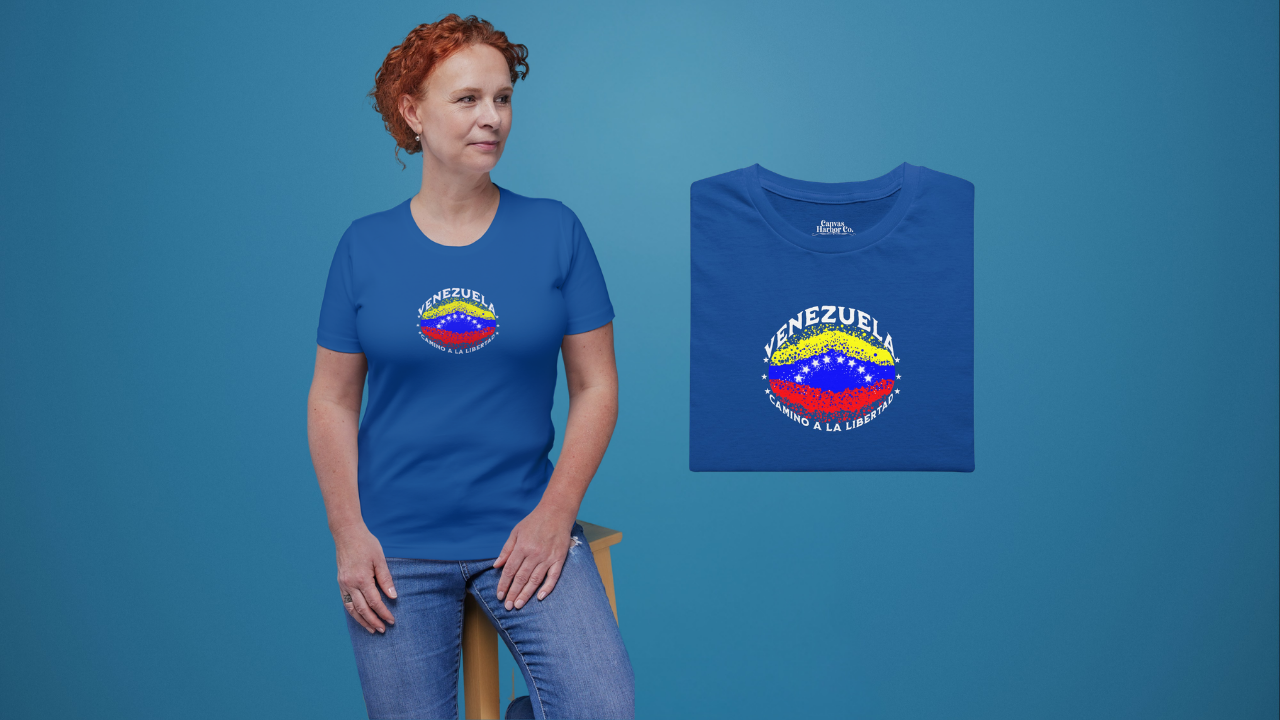 Franela azul, doblada, con una bandera venezolana de forma elíptica salpicada de puntos (como una explosión), con el nombre "VENEZUELA” arriba, el texto “CAMINO A LA LIBERTAD" abajo y 3 estrellas en cada lado.
A la izquierda, una mujer pelirroja viste la franela. Ella está sentada sobre un banco de madera, mirando a su izquierda.