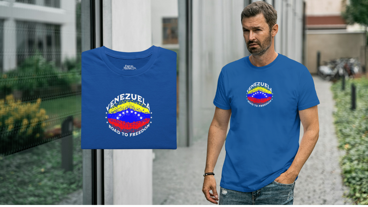 Blue folded T-shirt featuring an elliptical Venezuelan flag dotted with dots (like an explosion), with the word "VENEZUELA” at the top, the text “ROAD TO FREEDOM” below the flag, and 3 stars on each side.
On the right, a man wears the T-shirt. He’s standing, walking next to a building.