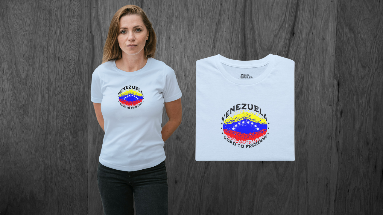 Light blue folded T-shirt featuring an elliptical Venezuelan flag dotted with dots (like an explosion), with the word "VENEZUELA” at the top, the text “ROAD TO FREEDOM” below the flag, and 3 stars on each side.
On the left, a blonde woman wears the T-shirt. She’s standing, facing forward, with both hands behind her back.