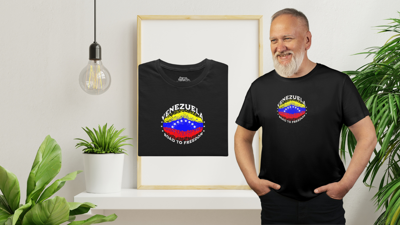 Black folded T-shirt featuring an elliptical Venezuelan flag dotted with dots (like an explosion), with the word "VENEZUELA” at the top, the text “ROAD TO FREEDOM” below the flag, and 3 stars on each side.
On the right, a bearded man wears the T-shirt. He’s standing, smiling, with both hands in his pants pockets.