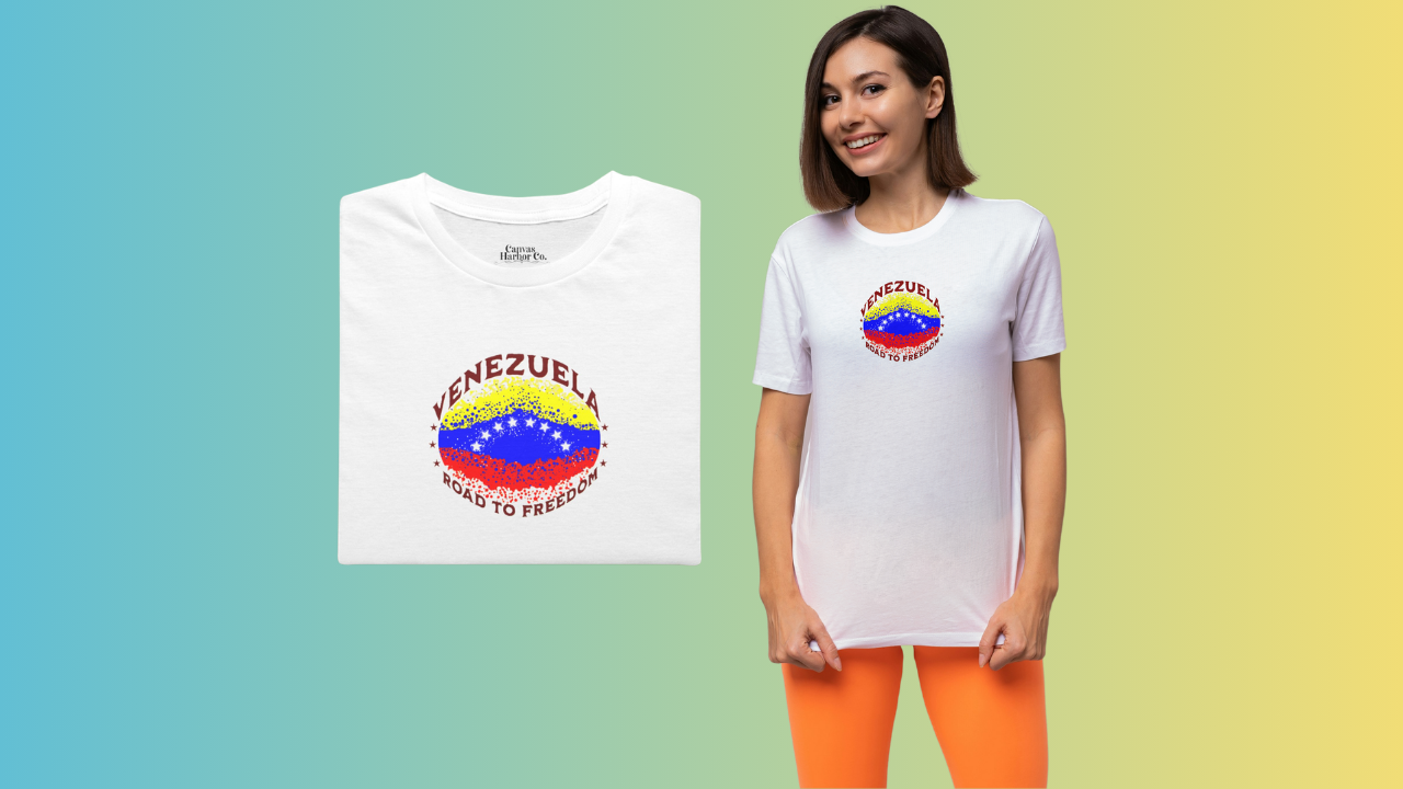 White folded T-shirt featuring an elliptical Venezuelan flag dotted with dots (like an explosion), with the word "VENEZUELA” at the top, the text “ROAD TO FREEDOM” below the flag, and 3 stars on each side.
On the right, a smiling woman wears the T-shirt. She’s standing, facing forward.
