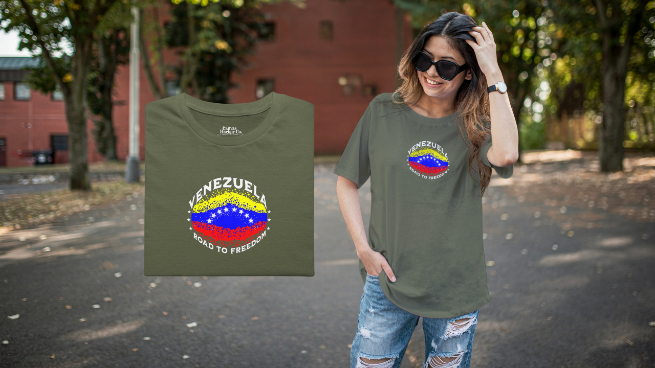 Olive green folded T-shirt featuring an elliptical Venezuelan flag dotted with dots (like an explosion), with the word "VENEZUELA” at the top, the text “ROAD TO FREEDOM” below the flag, and 3 stars on each side.
On the right, a young woman wears the T-shirt. She’s standing, smiling, and wearing sunglasses.