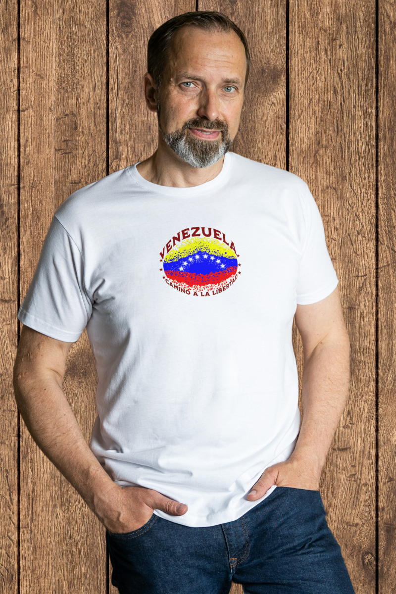 Un hombre viste una franela blanca con una bandera venezolana de forma elíptica salpicada de puntos (como una explosión), con el nombre "VENEZUELA” arriba, el texto “CAMINO A LA LIBERTAD" abajo y 3 estrellas en cada lado.
Él está de pie, mirando al frente, con ambas manos en los bolsillos de su pantalón.