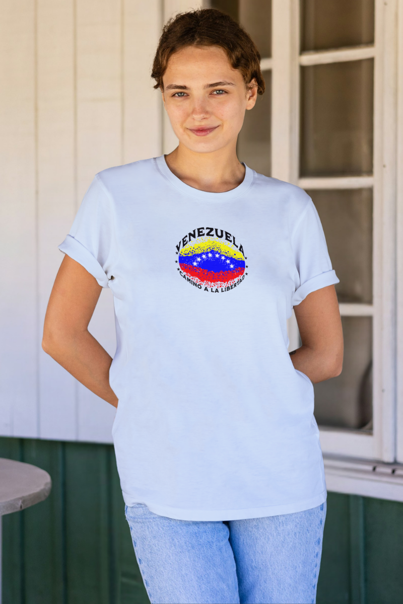 Una mujer joven viste una franela color azul claro con una bandera venezolana de forma elíptica salpicada de puntos (como una explosión), con el nombre "VENEZUELA” arriba, el texto “CAMINO A LA LIBERTAD" abajo y 3 estrellas en cada lado.
Ella sonríe, mirando al frente, con ambas manos detrás de su espalda.