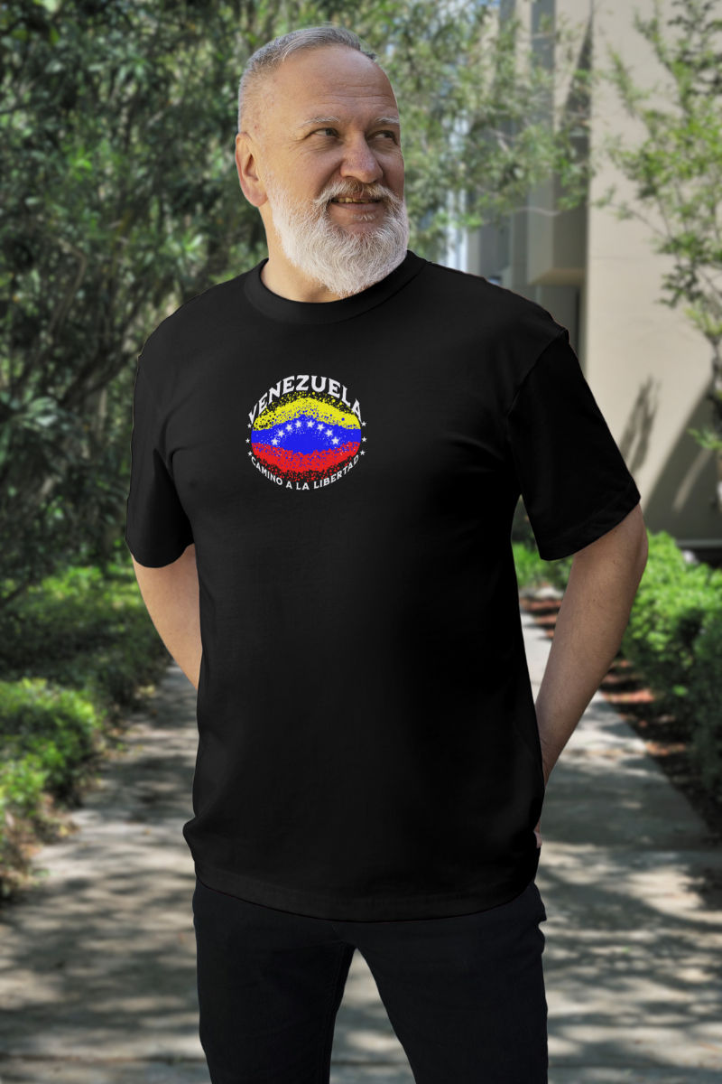 Un hombre con barba viste una franela negra con una bandera venezolana de forma elíptica salpicada de puntos (como una explosión), con el nombre "VENEZUELA” arriba, el texto “CAMINO A LA LIBERTAD" abajo y 3 estrellas en cada lado.
Él está de pie, mirando a su izquierda, con ambas manos detrás de su espalda.