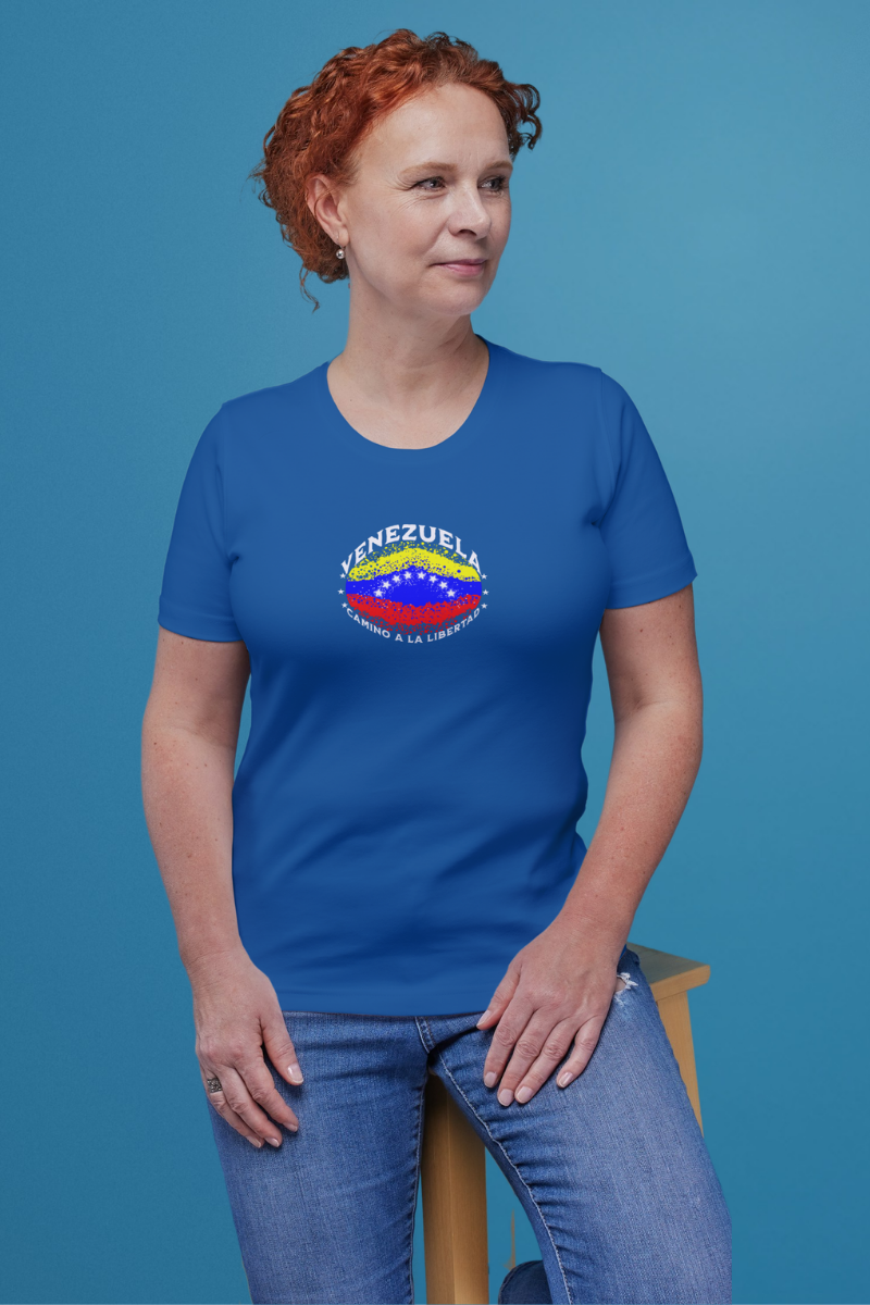 Una mujer pelirroja viste una franela azul con una bandera venezolana de forma elíptica salpicada de puntos (como una explosión), con el nombre "VENEZUELA” arriba, el texto “CAMINO A LA LIBERTAD" abajo y 3 estrellas en cada lado.
Ella está sentada sobre un banco de madera, mirando a su izquierda.