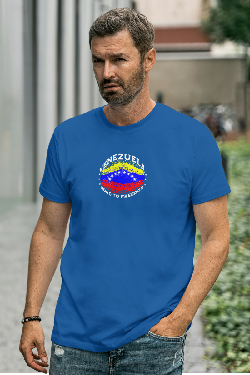 A man wears a blue T-shirt featuring an elliptical Venezuelan flag dotted with dots (like an explosion), with the word "VENEZUELA” at the top, the text “ROAD TO FREEDOM” below the flag, and 3 stars on each side.
He’s standing, walking next to a building.
