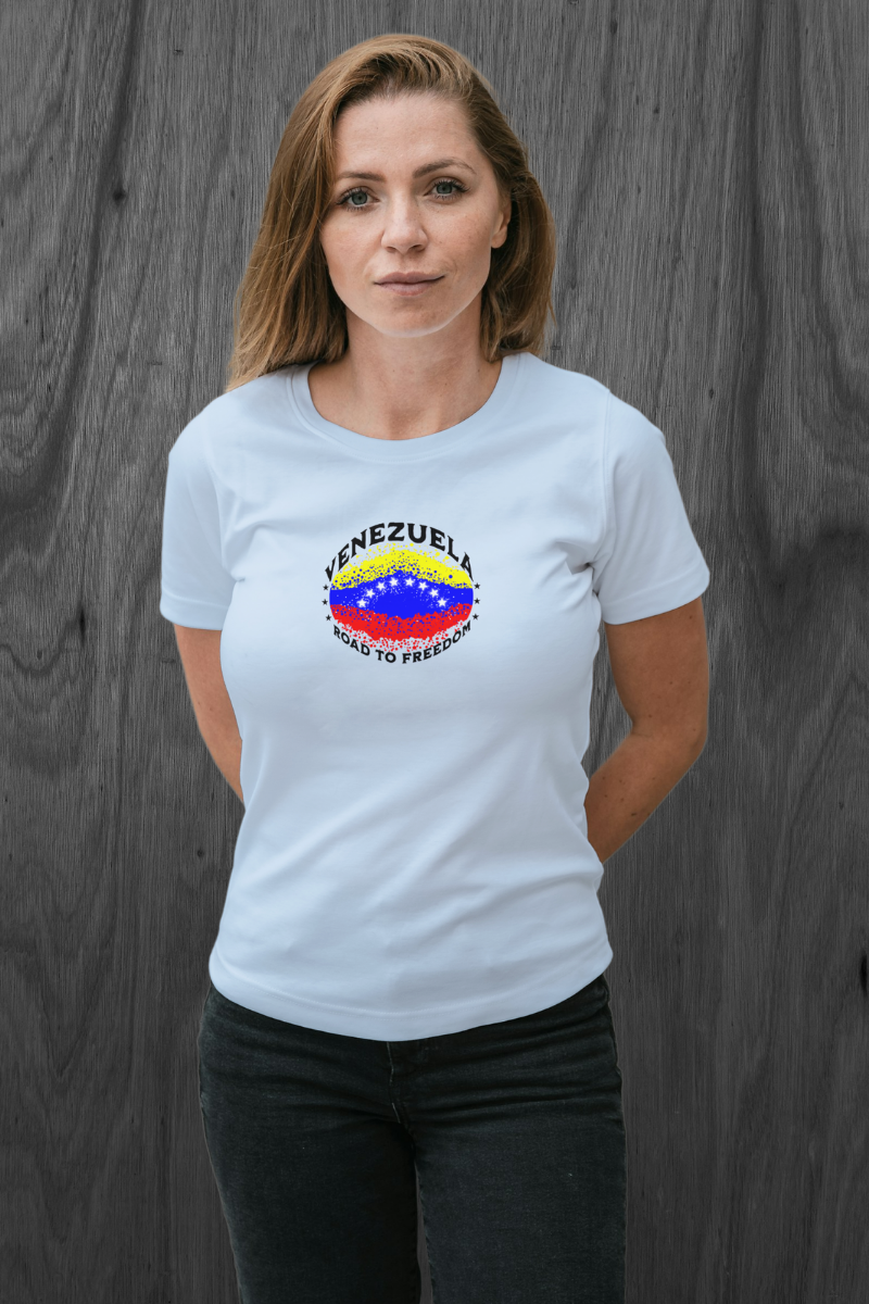 A blonde woman wears a light blue T-shirt featuring an elliptical Venezuelan flag dotted with dots (like an explosion), with the word "VENEZUELA” at the top, the text “ROAD TO FREEDOM” below the flag, and 3 stars on each side.
She’s standing, facing forward, with both hands behind her back.