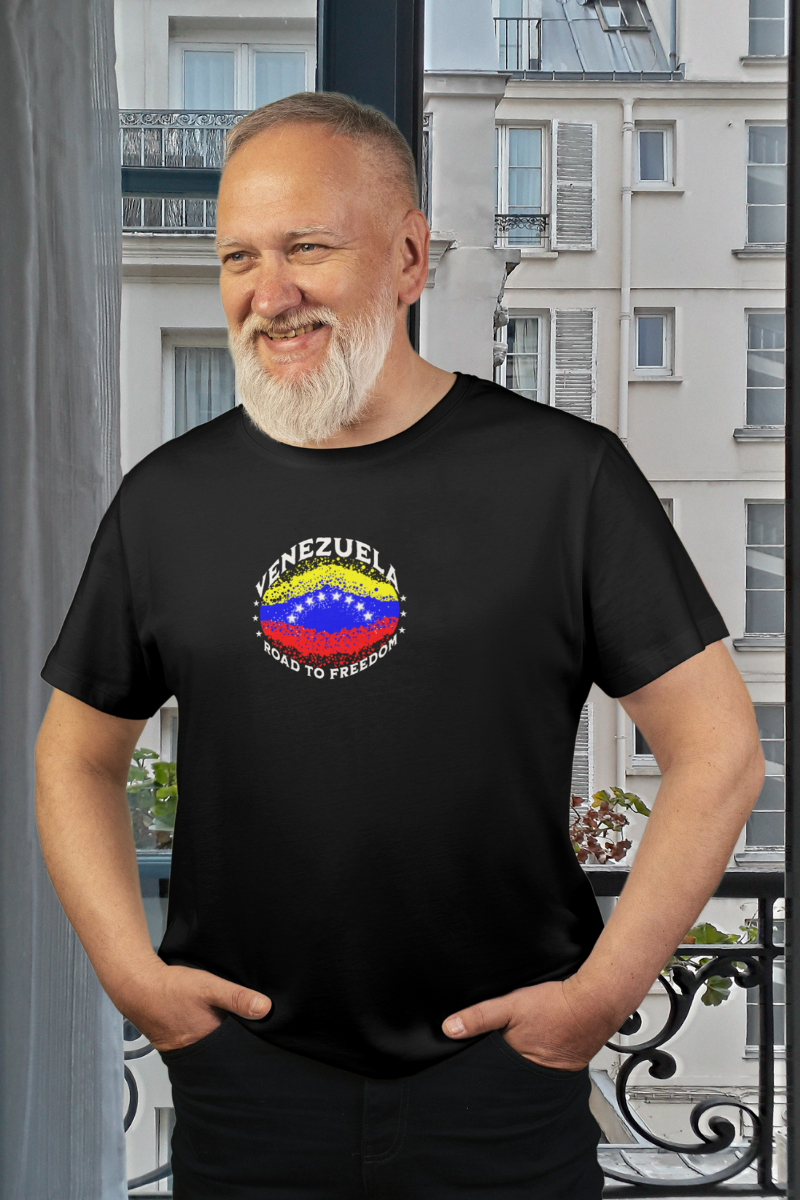 A bearded man wears a black T-shirt featuring an elliptical Venezuelan flag dotted with dots (like an explosion), with the word "VENEZUELA” at the top, the text “ROAD TO FREEDOM” below the flag, and 3 stars on each side.
He’s standing, smiling, with both hands in his pants pockets.
