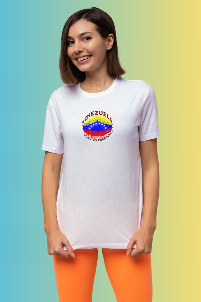 A smiling woman wears a white T-shirt featuring an elliptical Venezuelan flag dotted with dots (like an explosion), with the word "VENEZUELA” at the top, the text “ROAD TO FREEDOM” below the flag, and 3 stars on each side.
She’s standing, facing forward.