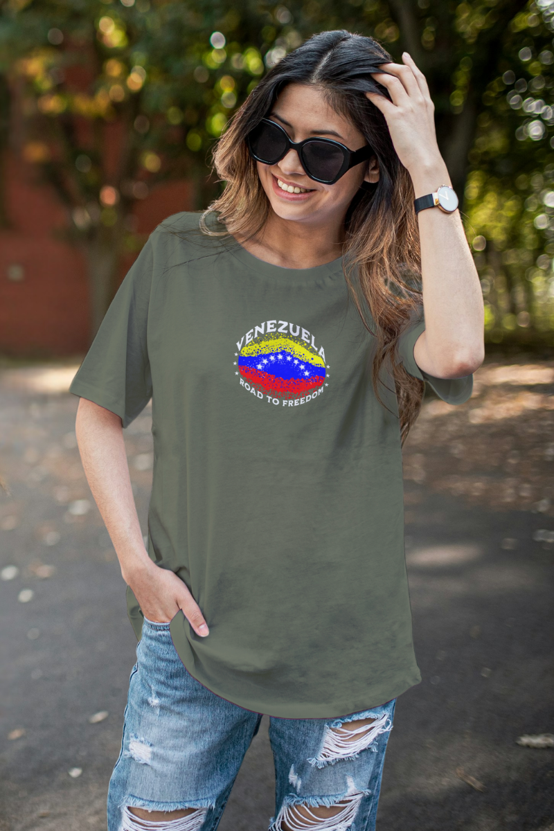 A young woman wears an olive-green T-shirt featuring an elliptical Venezuelan flag dotted with dots (like an explosion), with the word "VENEZUELA” at the top, the text “ROAD TO FREEDOM” below the flag, and 3 stars on each side.
She’s standing, smiling, and wearing sunglasses.