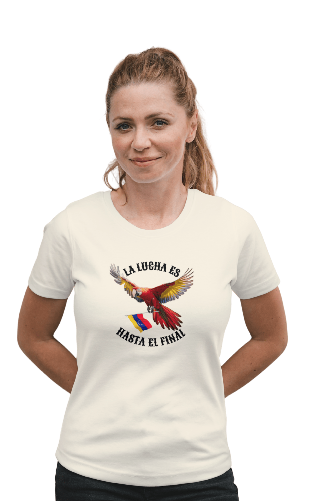 A blond woman is wearing a white unisex t-shirt featuring a colorful macaw flying while holding the Venezuelan flag. The text "La Lucha Es" is at the top, and "Hasta El Final" is below the macaw. She's smiling, both hands behind her back.
