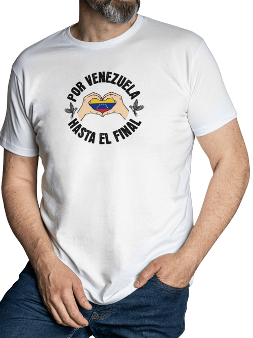 A man is wearing a white unisex t-shirt featuring a Venezuelan flag inside heart-shaped hands, the text "Por Venezuela Hasta El Final", and a pair of macaws on either side. He stands with his left hand in his pocket.