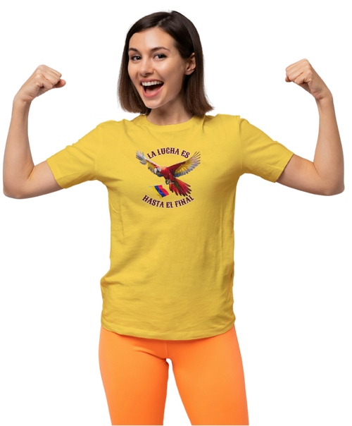 A woman is wearing a yellow unisex t-shirt featuring a colorful macaw flying while holding the Venezuelan flag. The text "La Lucha Es" is at the top, and "Hasta El Final" is below the macaw. She's smiling and flexing her arms in victory.